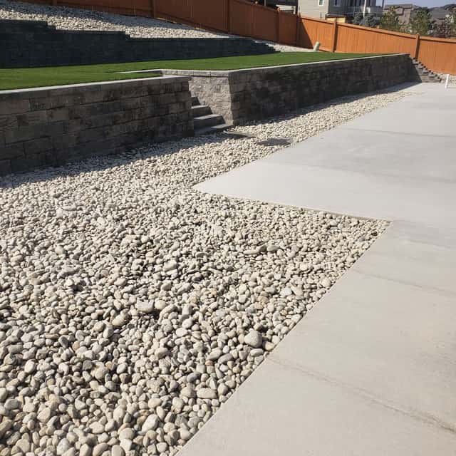 Broom-finished concrete patio beside a river-rock bed and block wall — Angel's Landscaping LLC, Castle Rock, CO